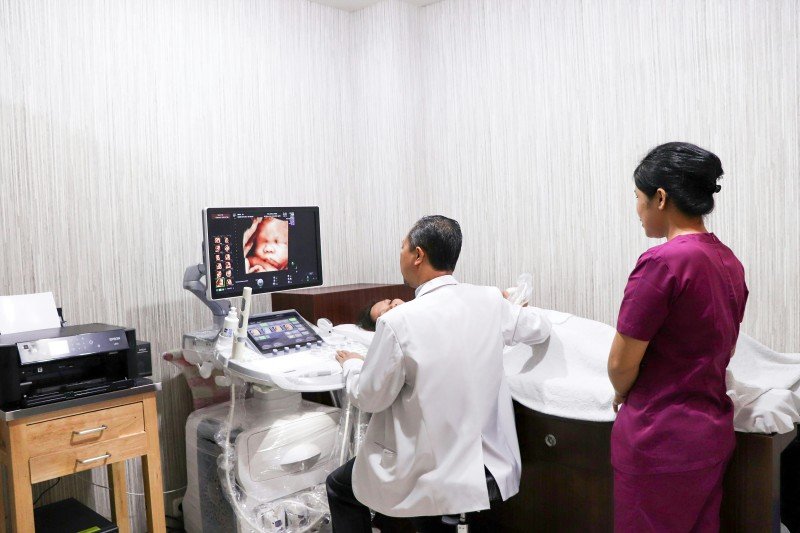 Doctor performing an ultrasound examination on a pregnant patient while a medical assistant stands nearby; the monitor displays a 3D image of the fetus.