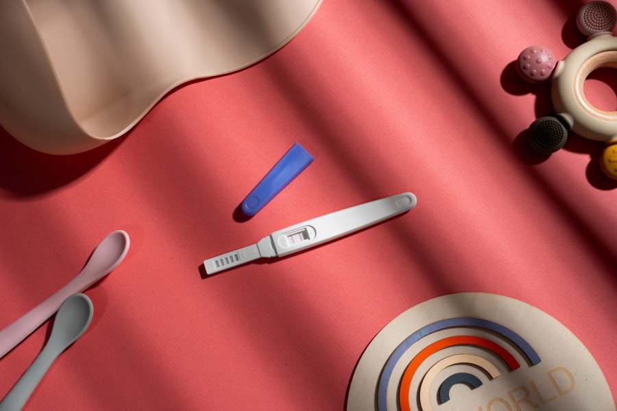 Positive pregnancy test lying on a pink surface, surrounded by baby items and decorative object